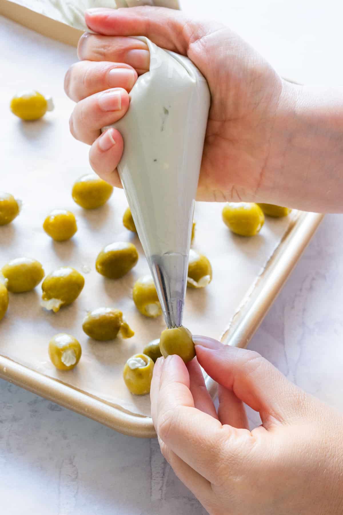 Hand using a piping bag to fill green olives with the cheese mixture on a parchment-lined tray.