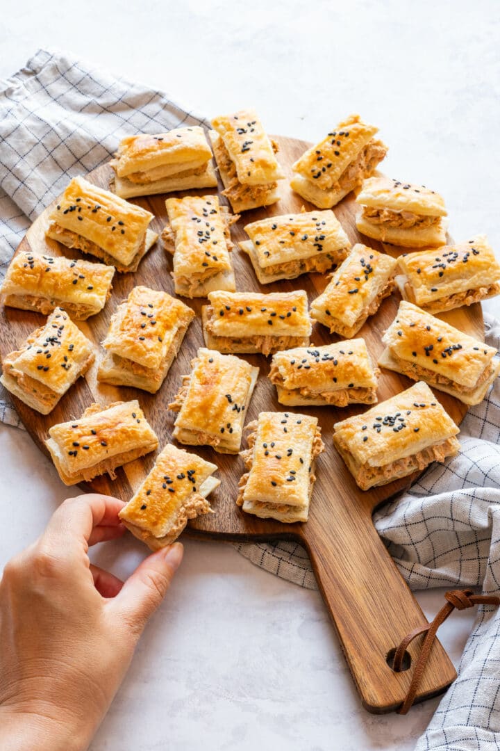 Hand reaching for one of the chicken puff pastries from a wooden serving board.