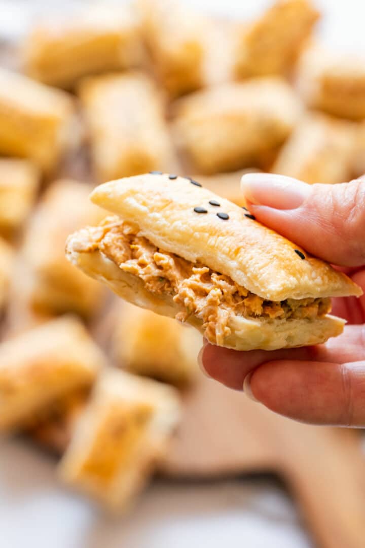 Close-up of a hand holding a sun-dried tomato chicken puff with creamy filling and flaky pastry topped with black sesame seeds.