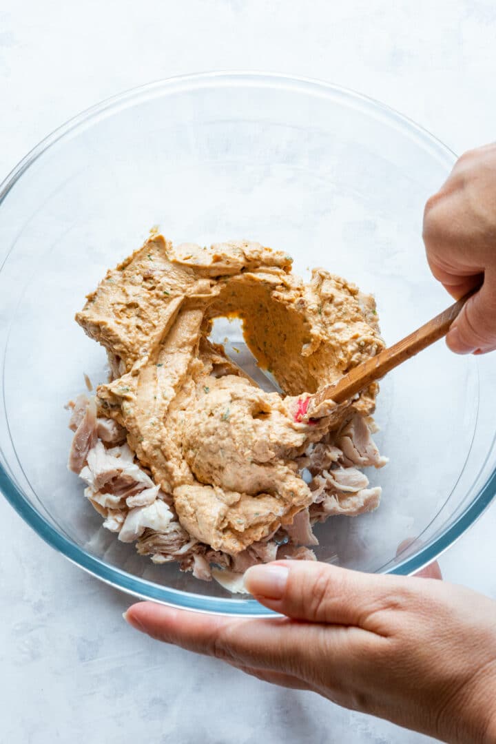 Chicken being mixed with the cream cheese and sun-dried tomato blend in a glass bowl.