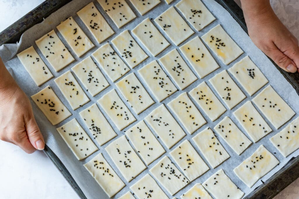 Unbaked puff pastry pieces sprinkled with black sesame seeds, arranged evenly on a baking sheet.
