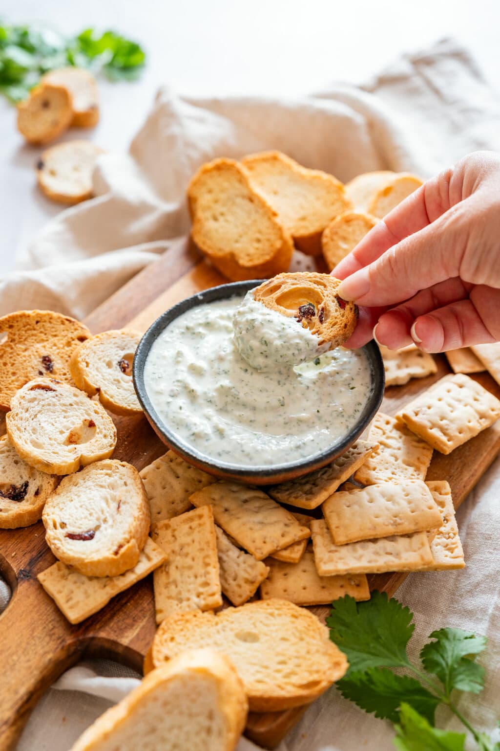 Creamy cilantro dip served in a small bowl, with a hand dipping toasted bread into it, surrounded by crackers and crostini