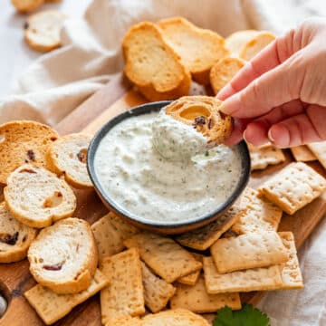 Creamy cilantro dip served in a small bowl, with a hand dipping toasted bread into it, surrounded by crackers and crostini
