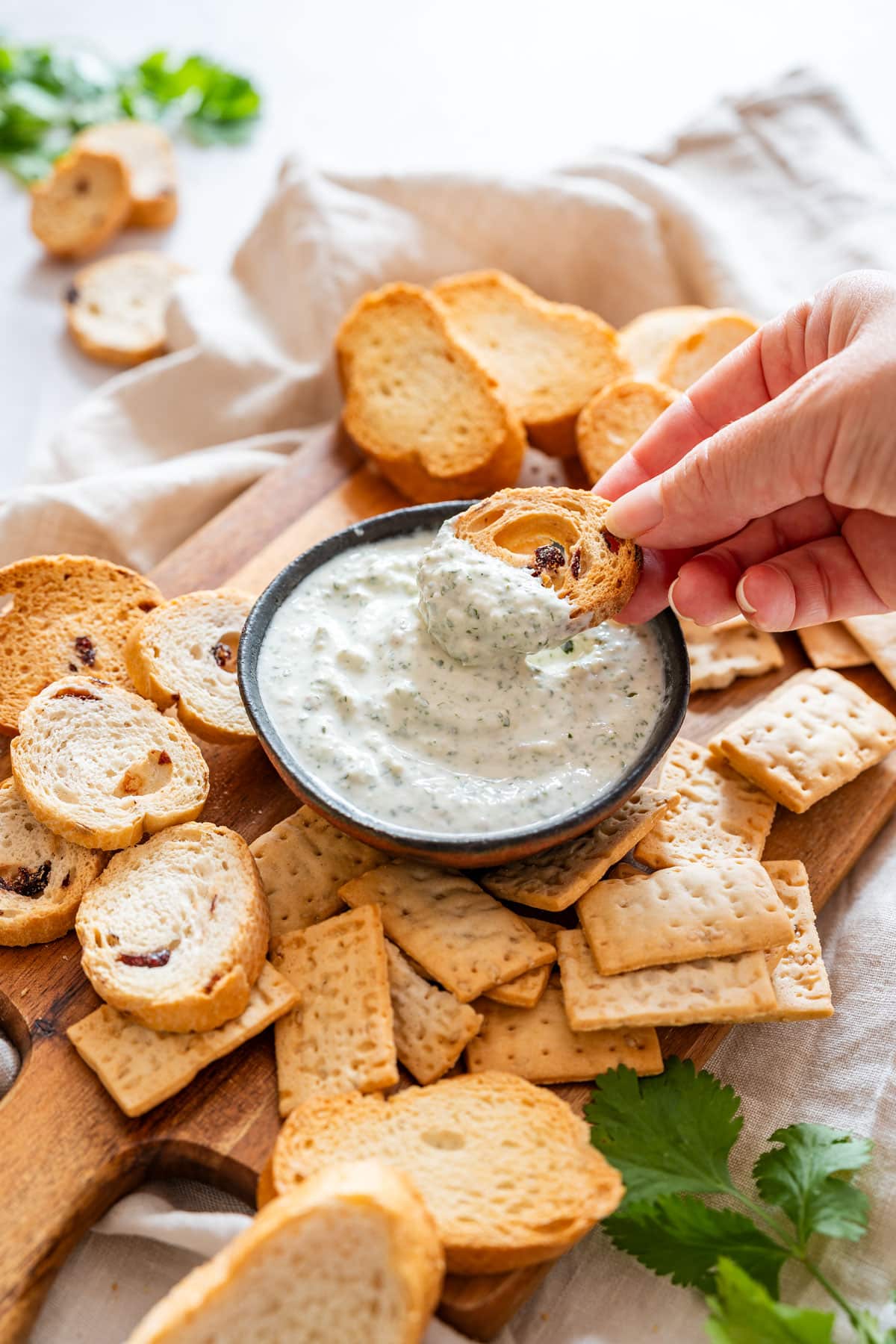 Creamy cilantro dip served in a small bowl, with a hand dipping toasted bread into it, surrounded by crackers and crostini