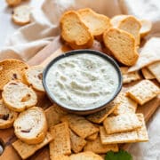 Cilantro yogurt dip in a black bowl surrounded by crackers, toast rounds, and a sprig of fresh cilantro.