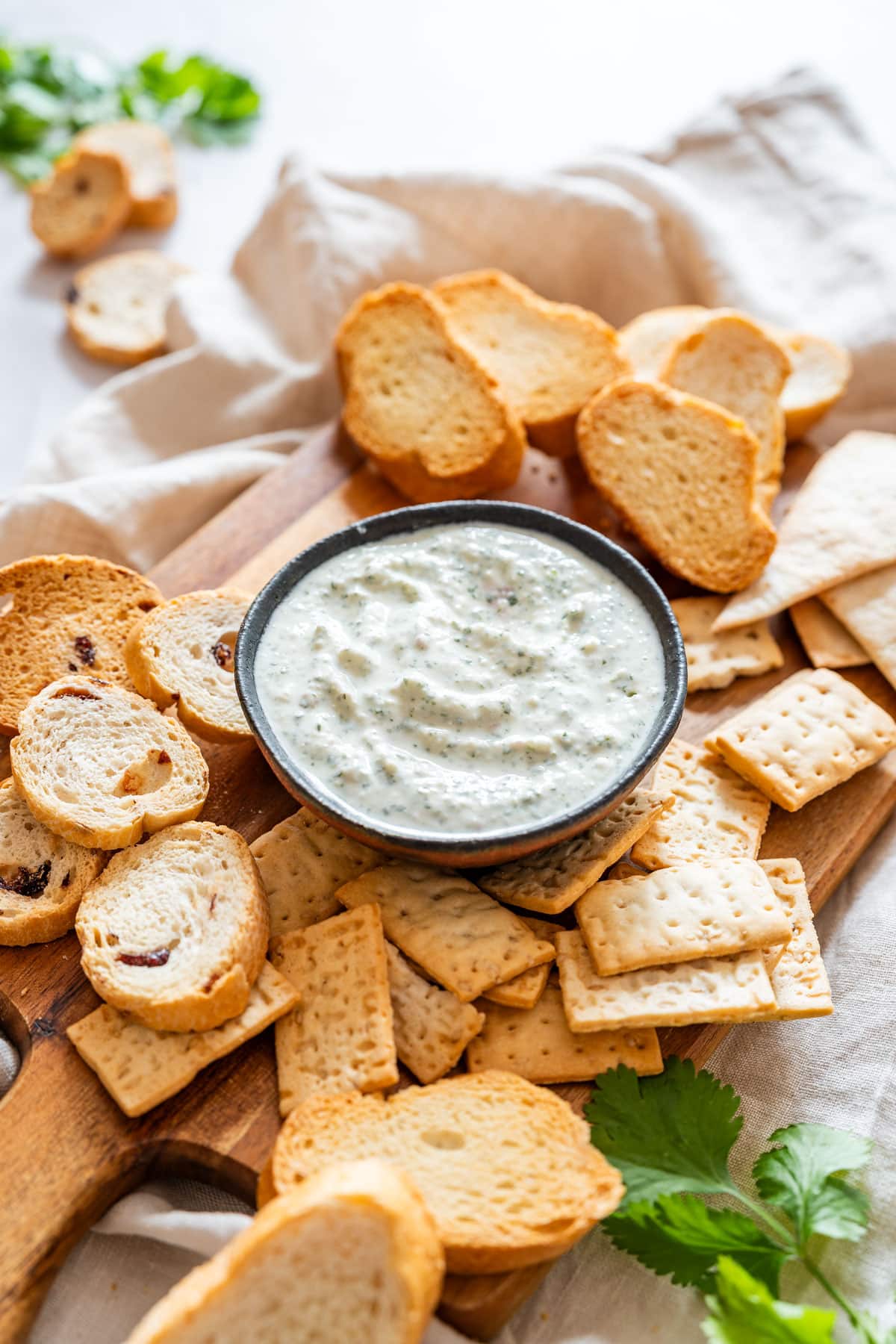 Cilantro yogurt dip in a black bowl surrounded by crackers, toast rounds, and a sprig of fresh cilantro.