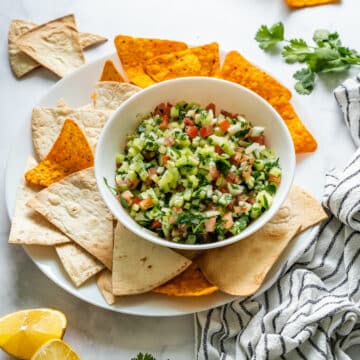 Bowl of fresh cucumber salsa served with tortilla chips and flatbread triangles on a white plate.