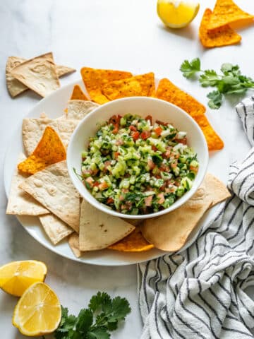 Bowl of fresh cucumber salsa served with tortilla chips and flatbread triangles on a white plate.