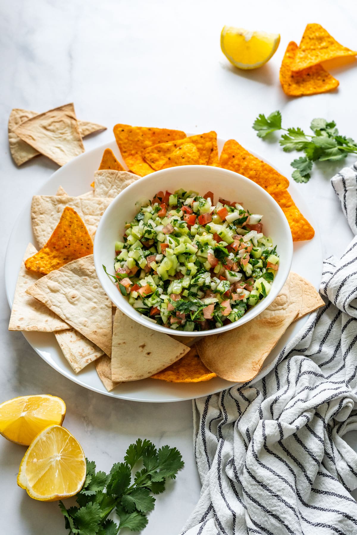 Bowl of fresh cucumber salsa served with tortilla chips and flatbread triangles on a white plate.