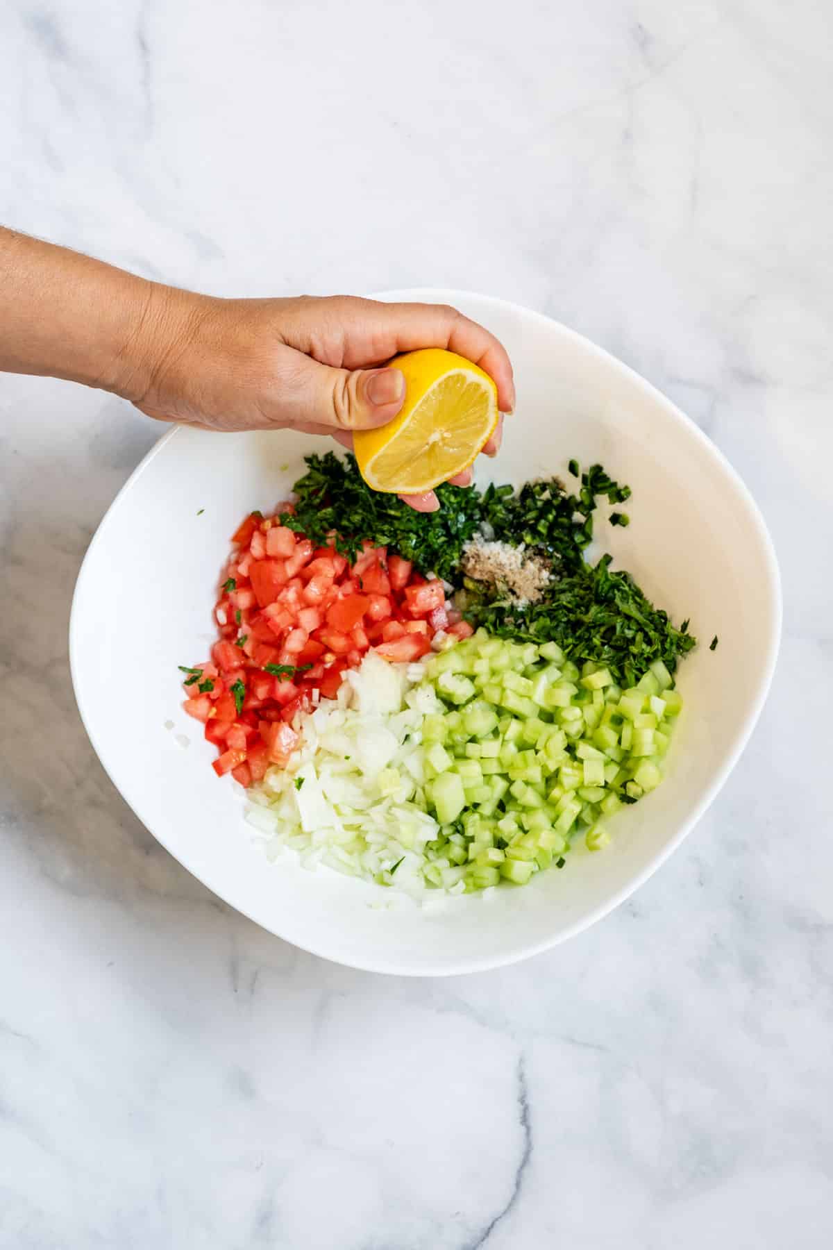 Lemon being squeezed over chopped vegetables in a white mixing bowl.