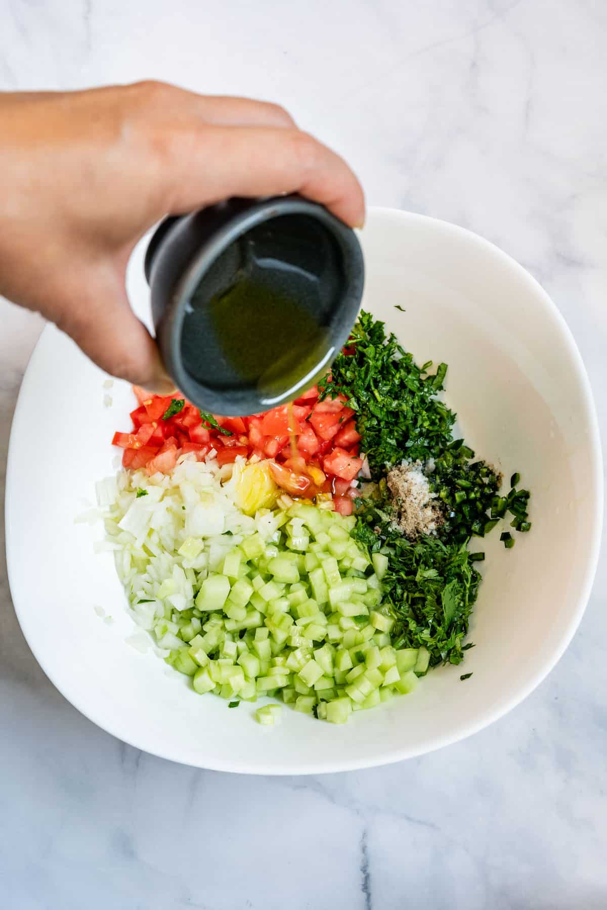 Olive oil being poured into a bowl of diced cucumber, tomato, onion, herbs and spices.