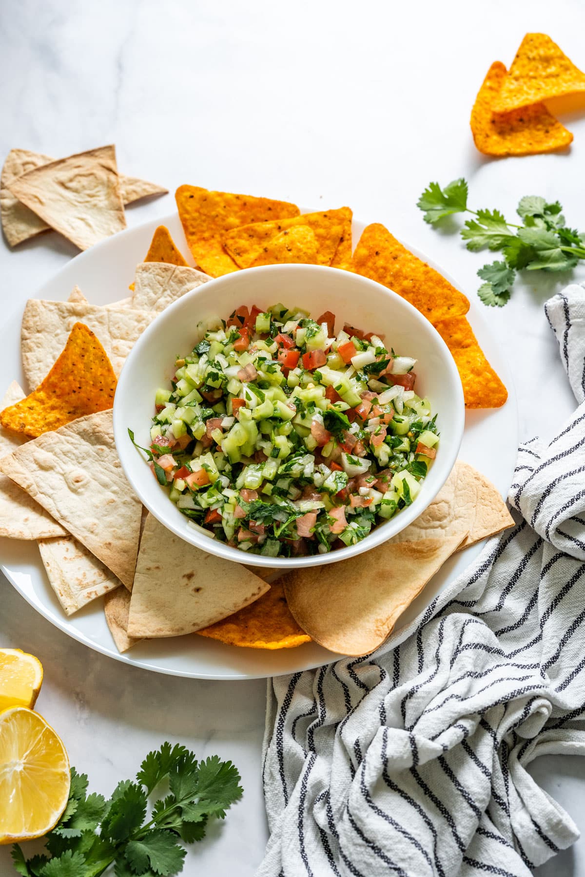 Prepared cucumber salsa in a white bowl, surrounded by chips and flatbread on a plate.