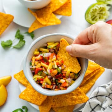 A hand dipping a tortilla chip into a bowl of peach salsa.