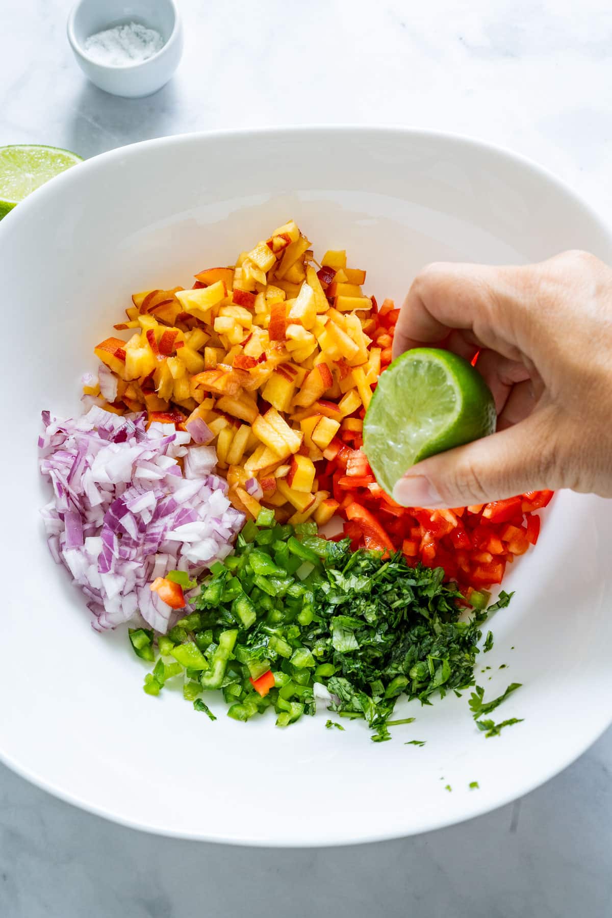 Lime being squeezed over chopped peaches, onions and peppers in a mixing bowl.