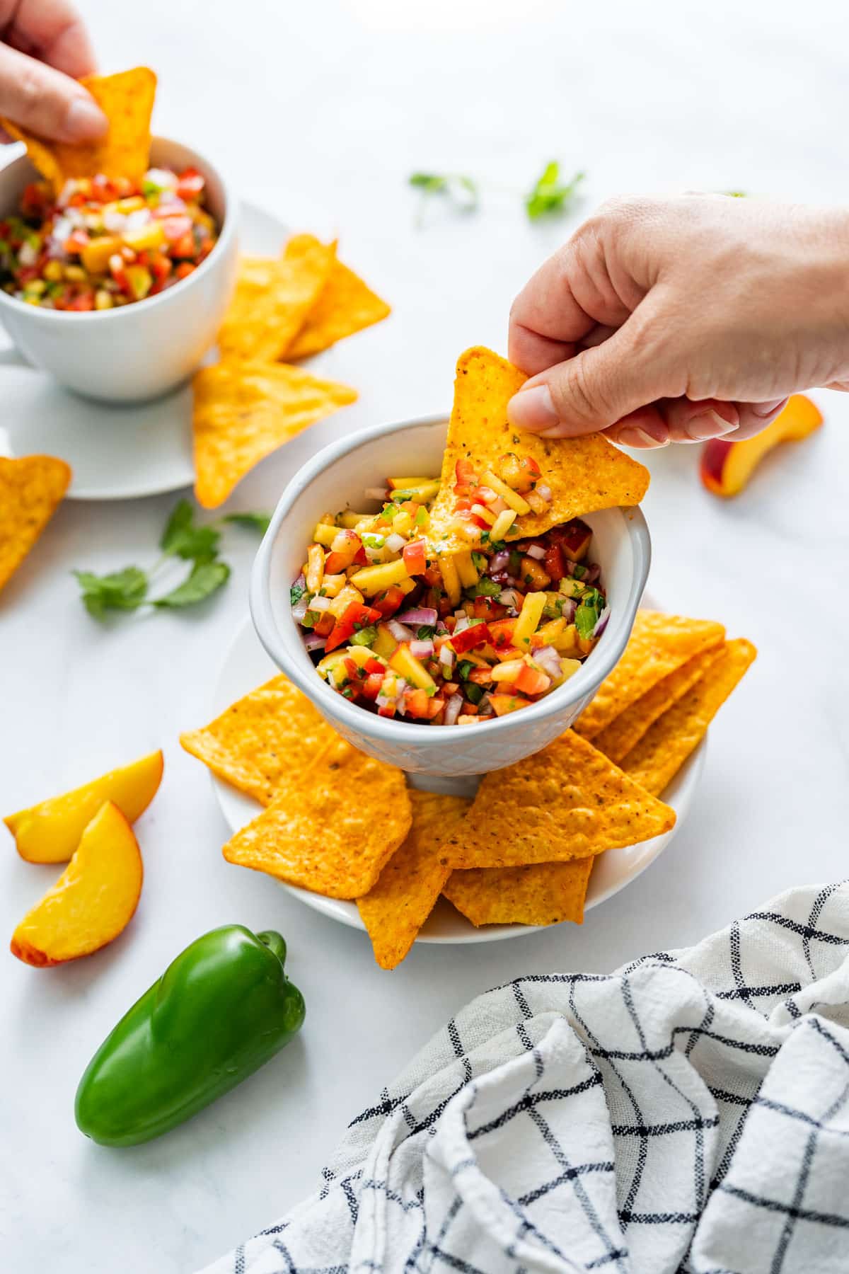 Hands dipping chips into fresh peach salsa with sliced peaches and a bell pepper in the background.
