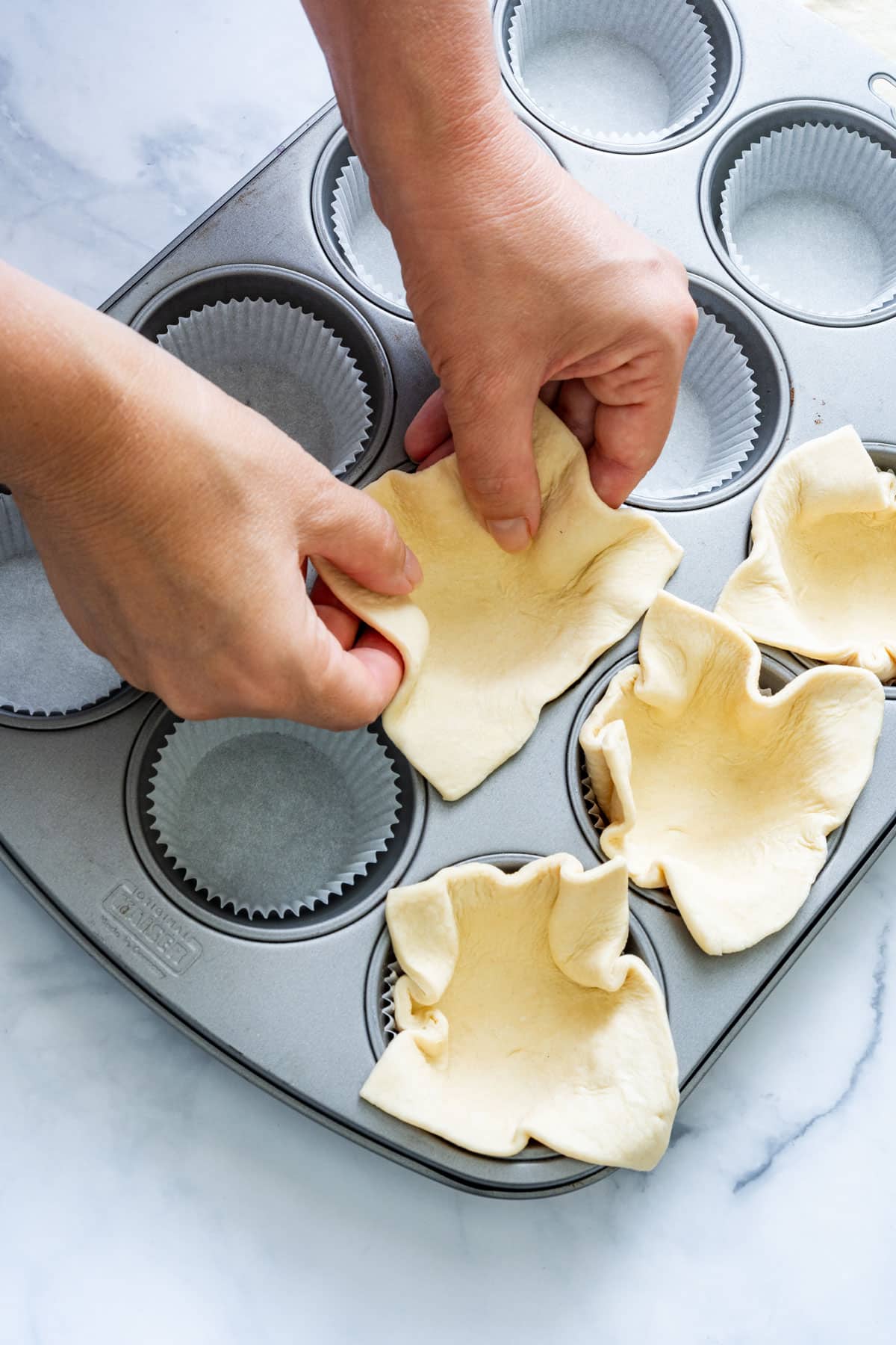 Hands pressing puff pastry squares into muffin tin cups.