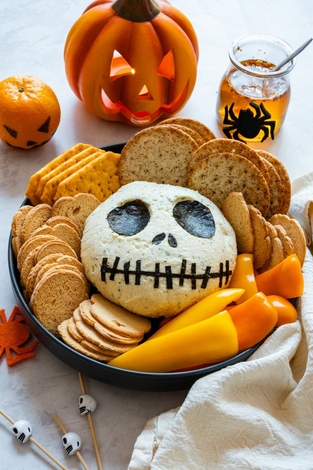 Jack Skellington cheese ball on a Halloween platter with crackers and peppers, lit jack-o’-lantern behind.
