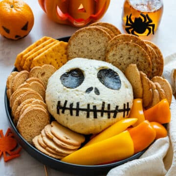 Jack Skellington cheese ball on a Halloween platter with crackers and peppers, lit jack-o’-lantern behind.