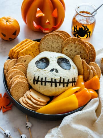 Jack Skellington cheese ball on a Halloween platter with crackers and peppers, lit jack-o’-lantern behind.