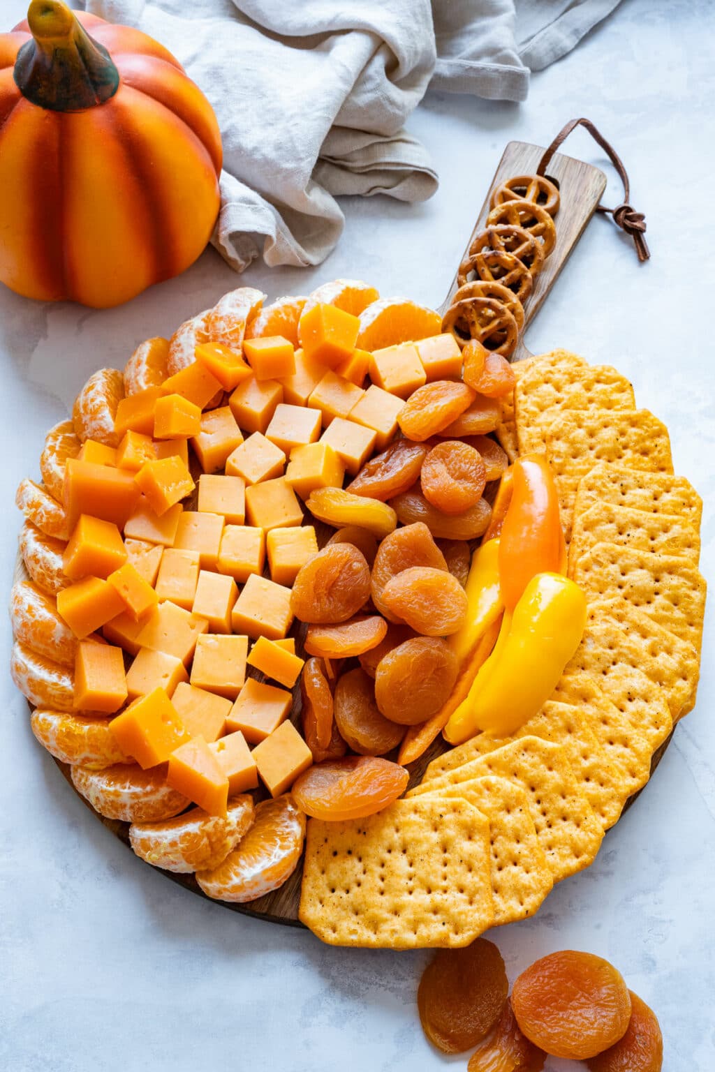 Pumpkin-shaped cheese board with cheddar cubes, mandarins, dried apricots, peppers, crackers, pretzel handle.