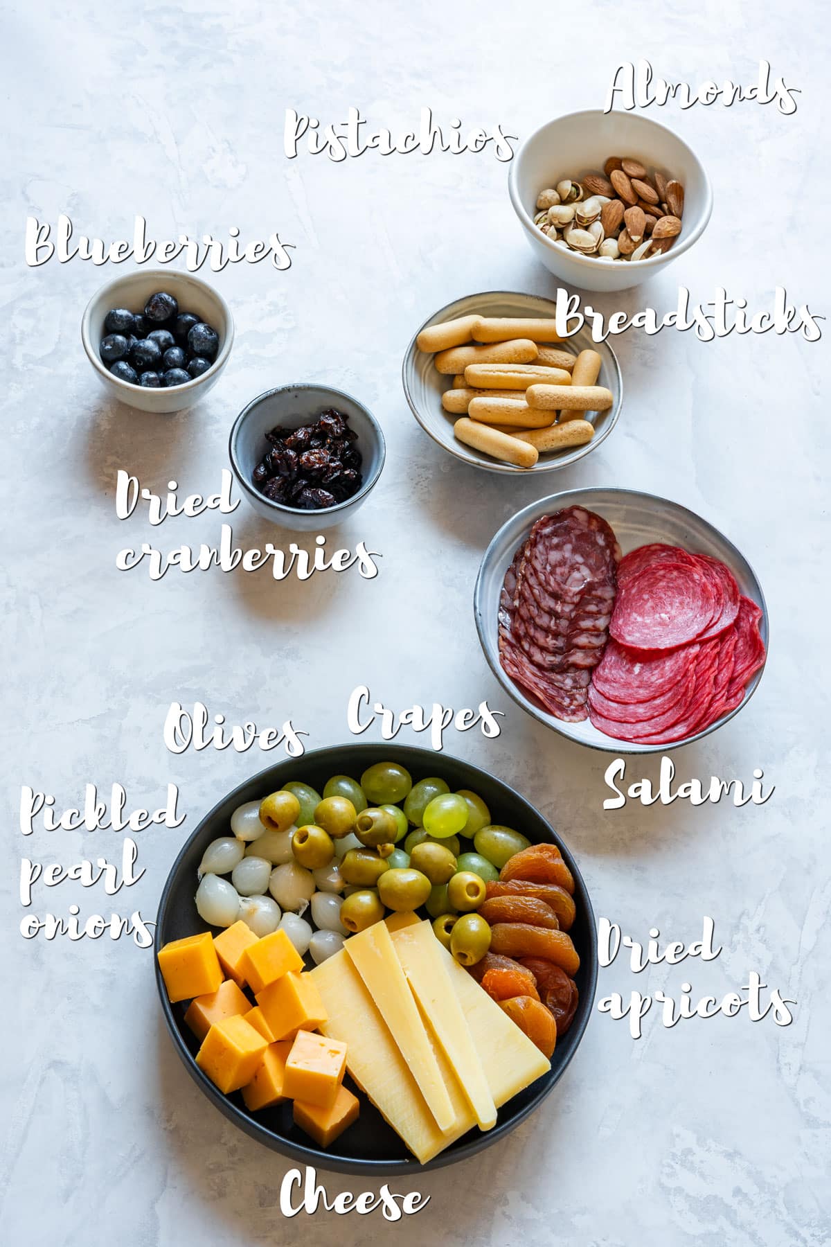 Charcuterie cup ingredients on a countertop: cheese, salami, grapes, olives, nuts, dried fruit, blueberries and breadsticks.