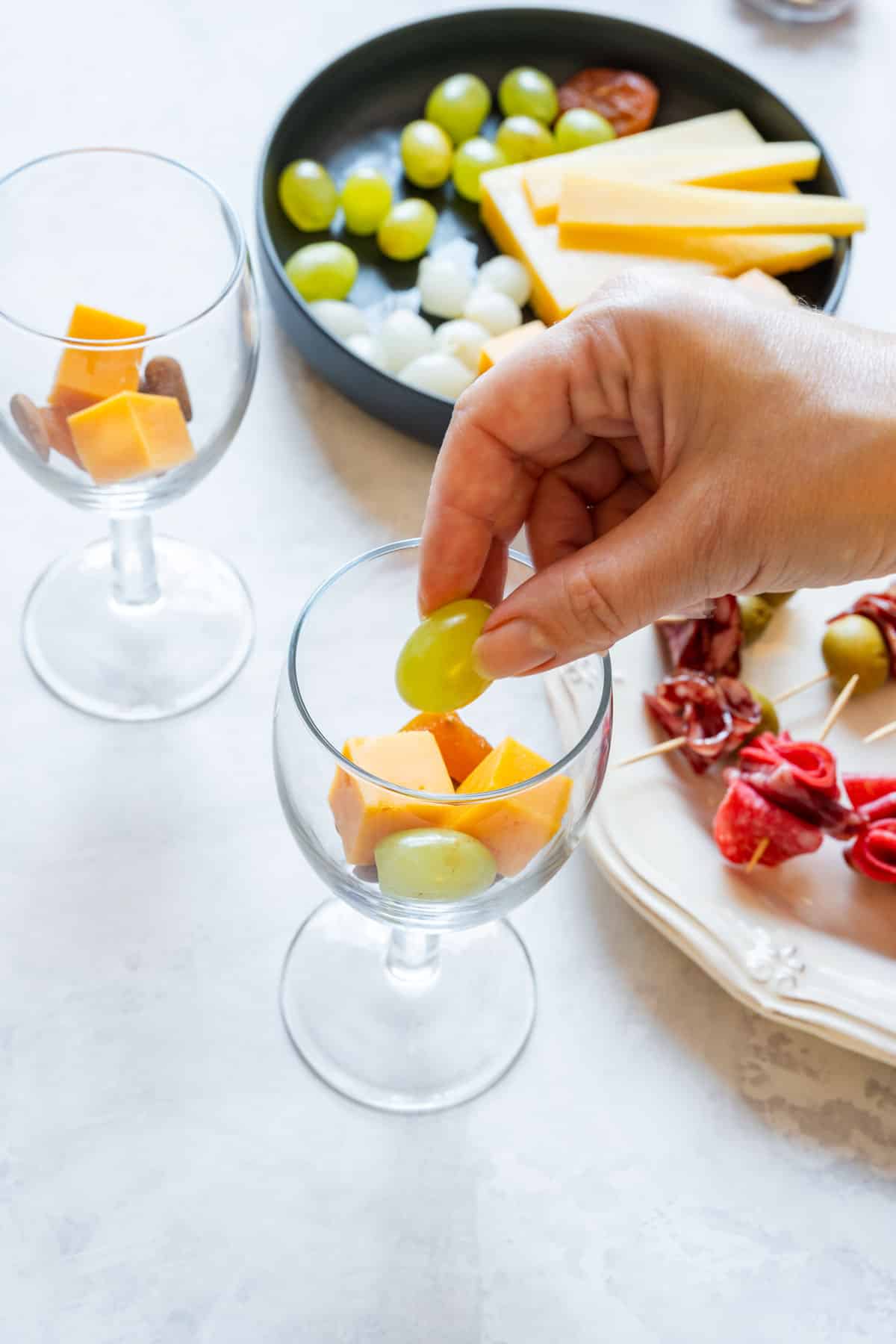 Adding a green grape to a glass with cheddar cubes while assembling individual snack cups.