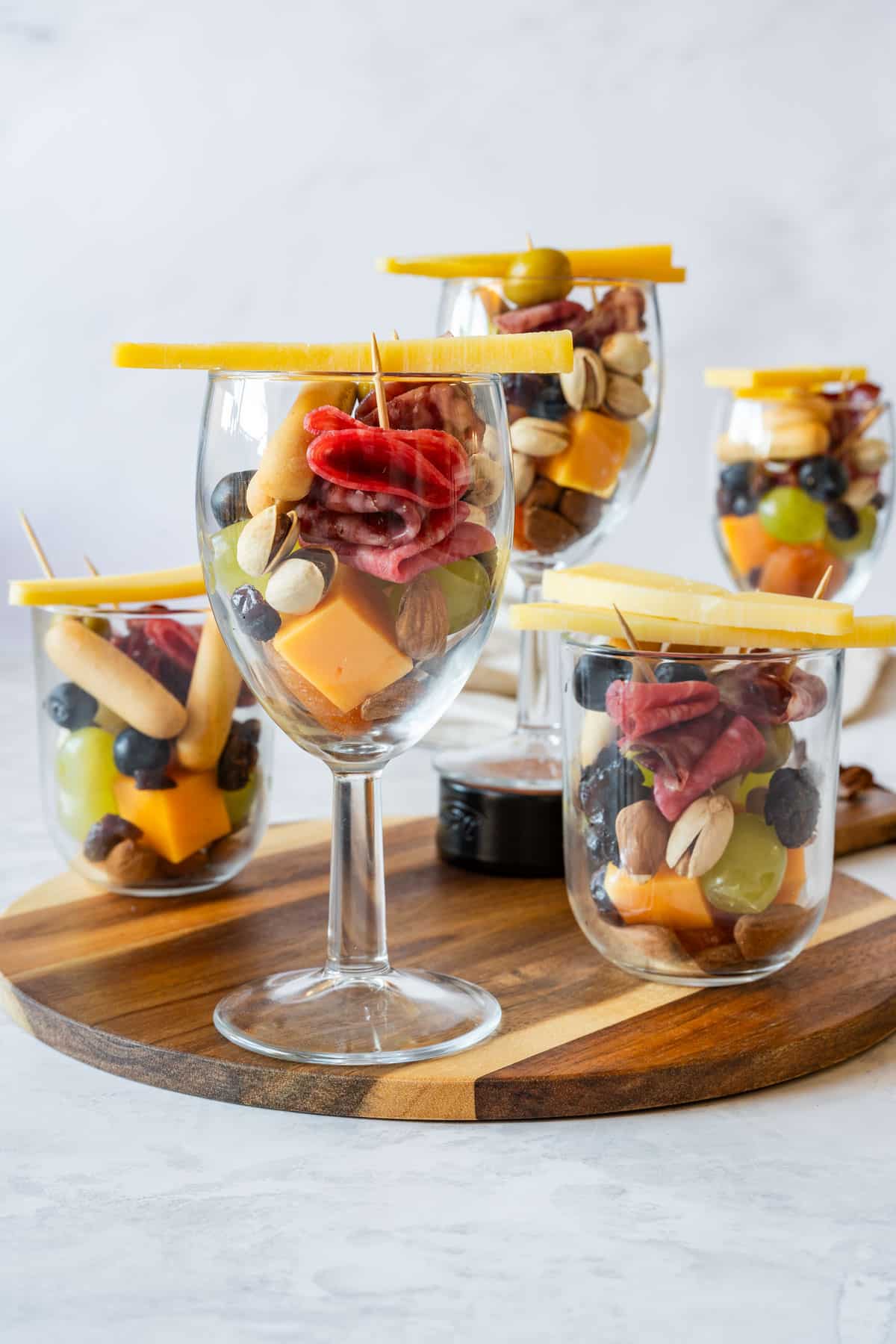 Assorted appetizer cups in mixed glassware with meat, cheese, fruit, nuts and cheese sticks on a wooden board.