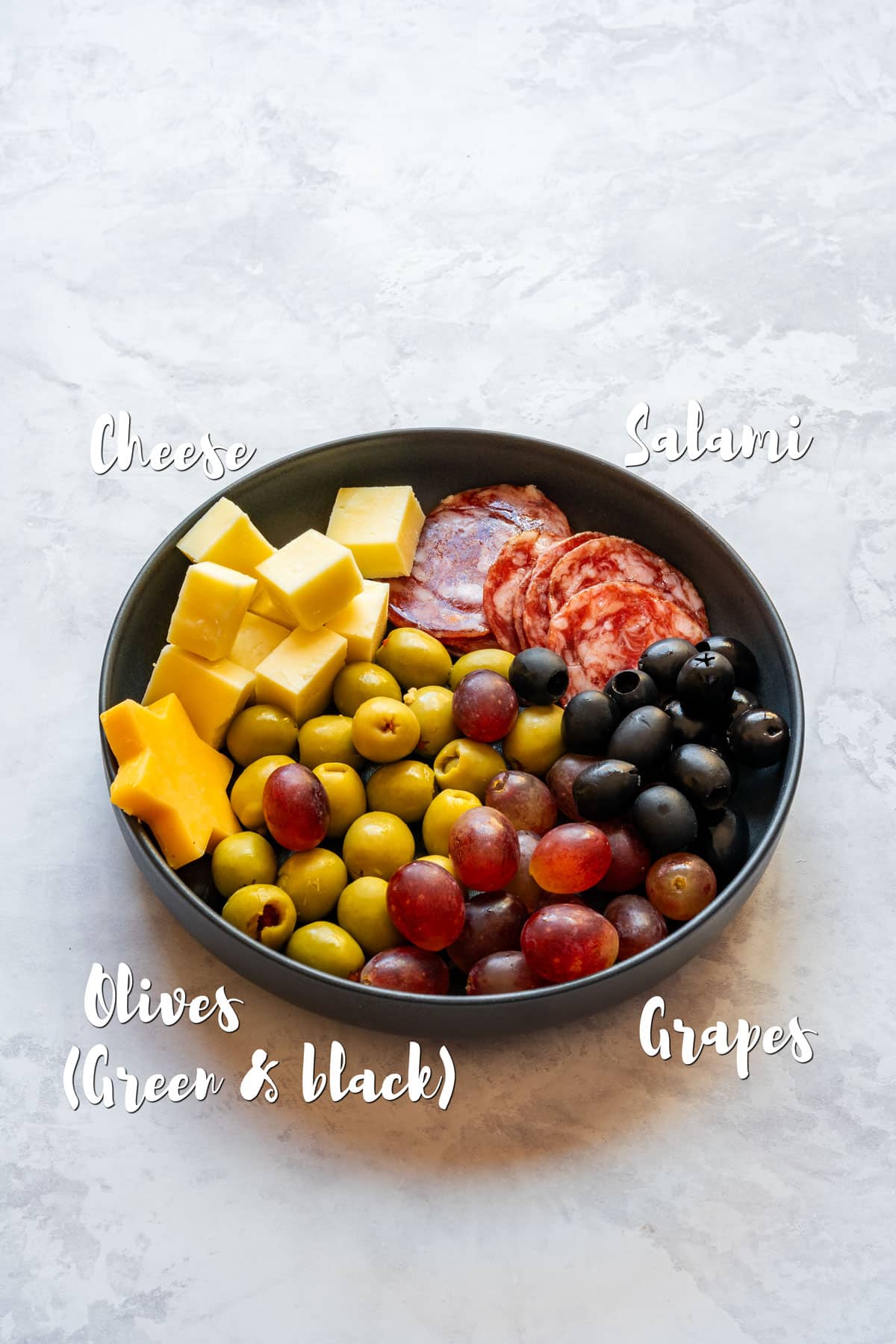 Prepared ingredients for the tree including cheese cubes and a cheese star, salami slices, green and black olives and grapes in a shallow bowl.