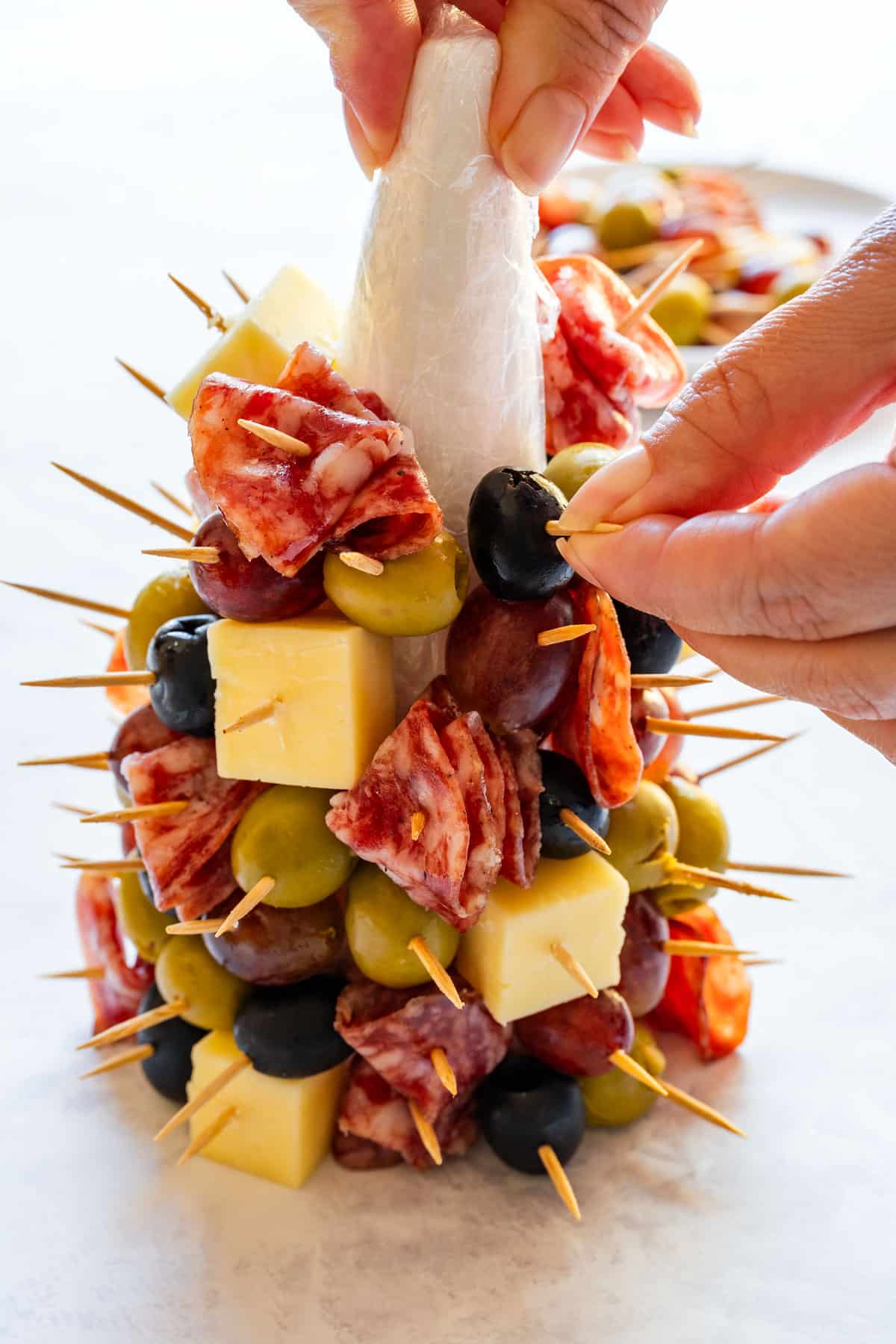 Building the tree by inserting toothpicks with cheese, salami, olives and grapes into the wrapped cone.