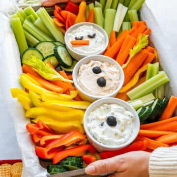 Hands holding a snowman veggie tray filled with carrots, celery, bell peppers, cucumber and three dips, with crackers on the side.