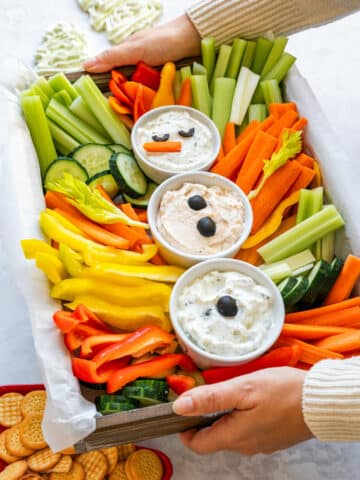 Hands holding a snowman veggie tray filled with carrots, celery, bell peppers, cucumber and three dips, with crackers on the side.