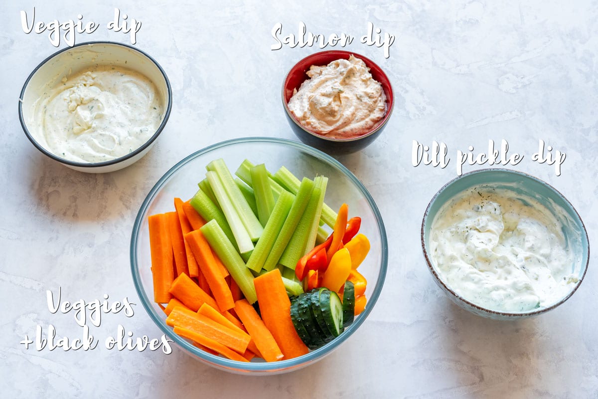 Veggie dip, salmon dip, dill pickle dip and a bowl of cut vegetables and black olives arranged as ingredients for a snowman veggie tray.