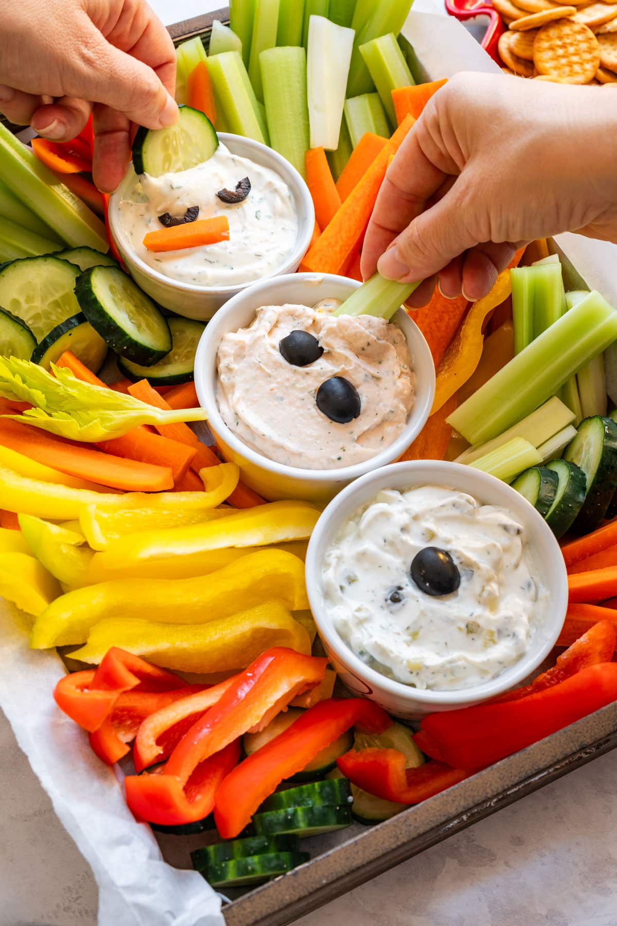 Two hands dipping cucumber and celery sticks into the snowman veggie tray dips surrounded by colorful fresh vegetables.