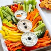 Finished snowman vegetable platter with three dips, celery and carrot sticks, bell peppers and cucumber with crackers in the background.