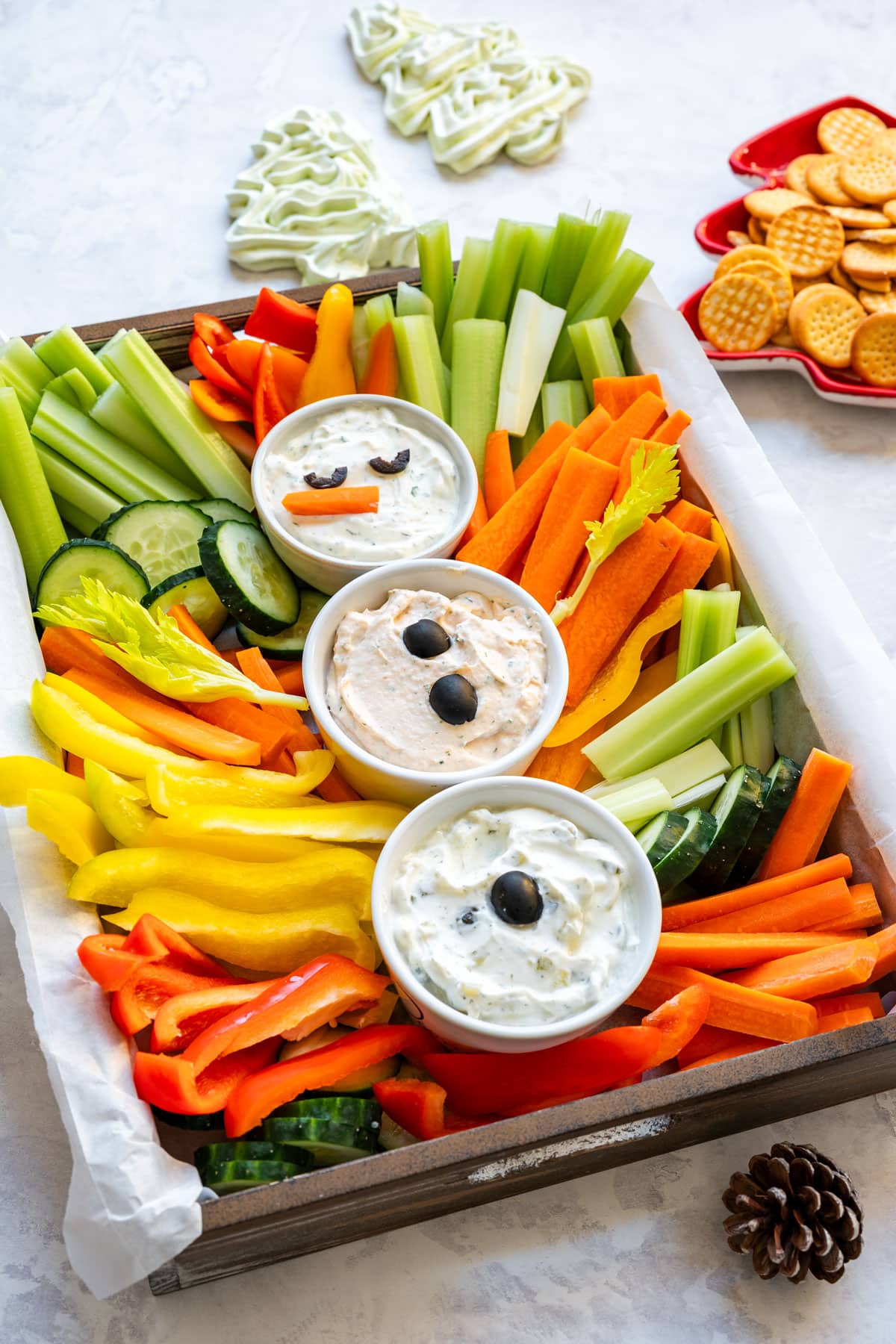 Finished snowman vegetable platter with three dips, celery and carrot sticks, bell peppers and cucumber with crackers in the background.