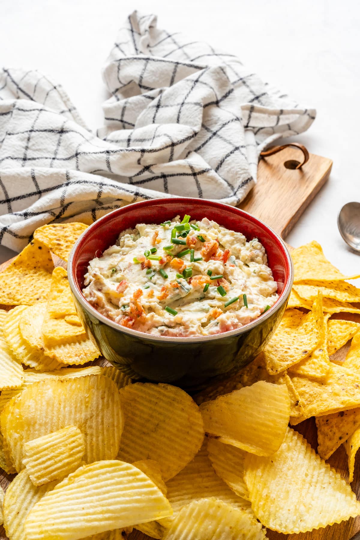 Loaded baked potato dip served with ruffled potato chips and tortilla chips on a wooden board.