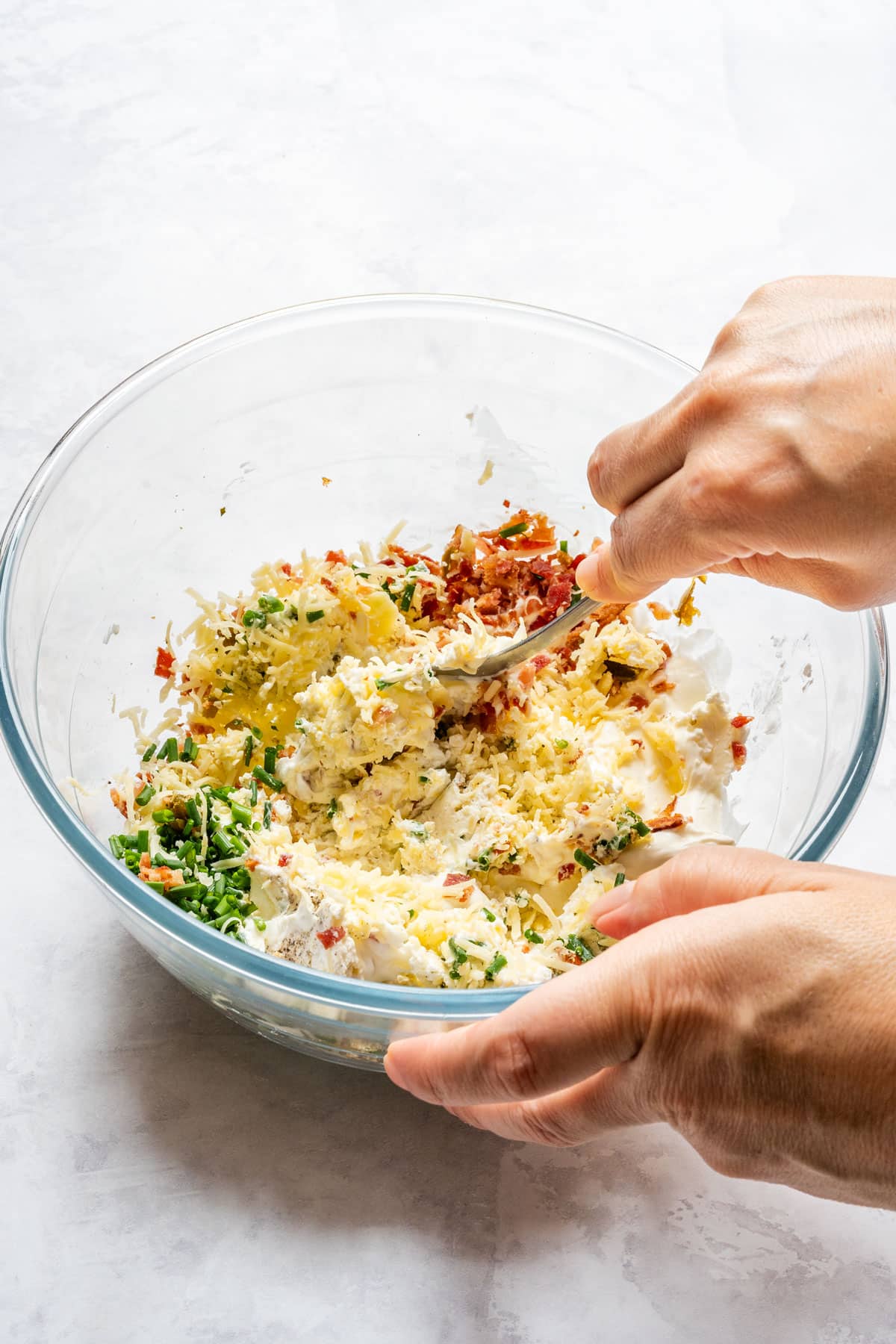 Hand mixing cream cheese, sour cream, cheddar, bacon, chives and jalapeños in a glass bowl.