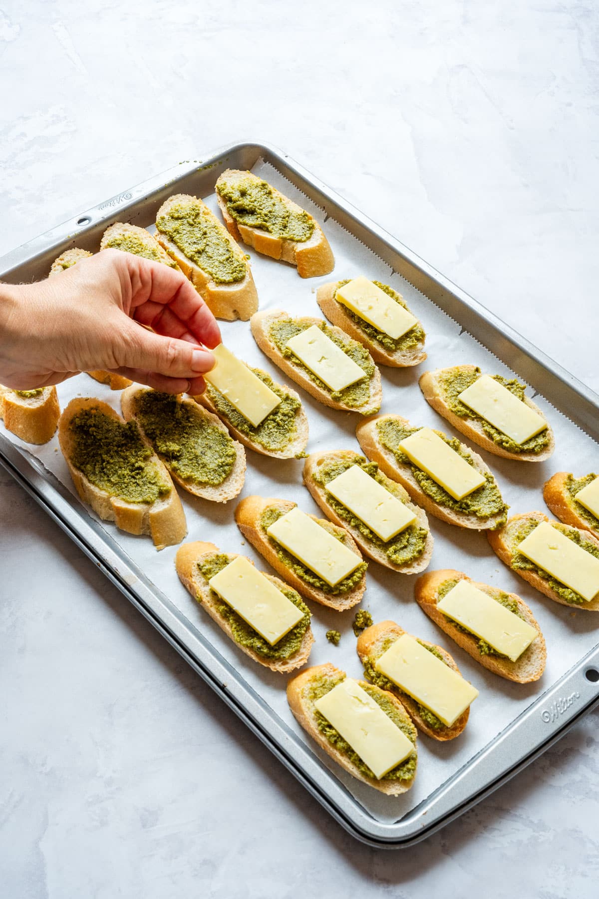 Hand placing cheddar cheese slices on baguette rounds spread with pesto.