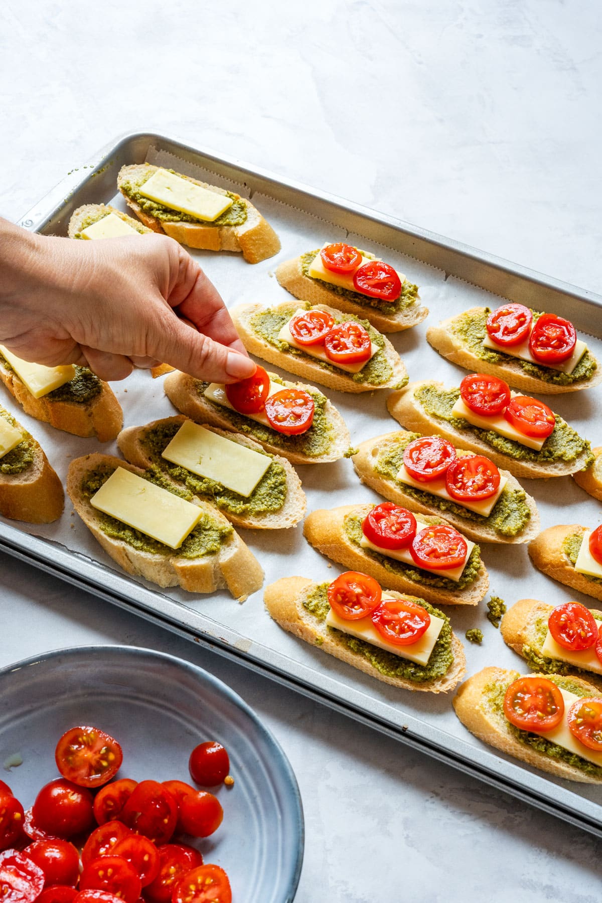 Hand adding cherry tomato slices on top of cheese-topped crostini before baking.