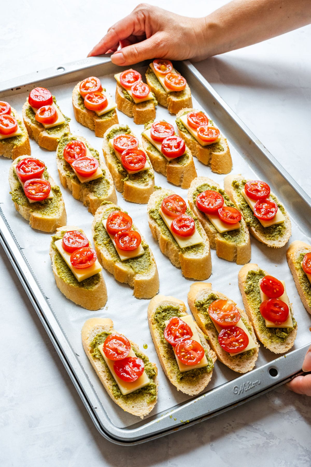 Unbaked crostini topped with pesto, cheddar cheese and cherry tomatoes on a sheet pan.