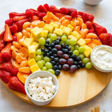 Rainbow fruit platter with fruit dip, mini marshmallows, strawberries, mandarins, pineapple, grapes and blueberries on a round board.