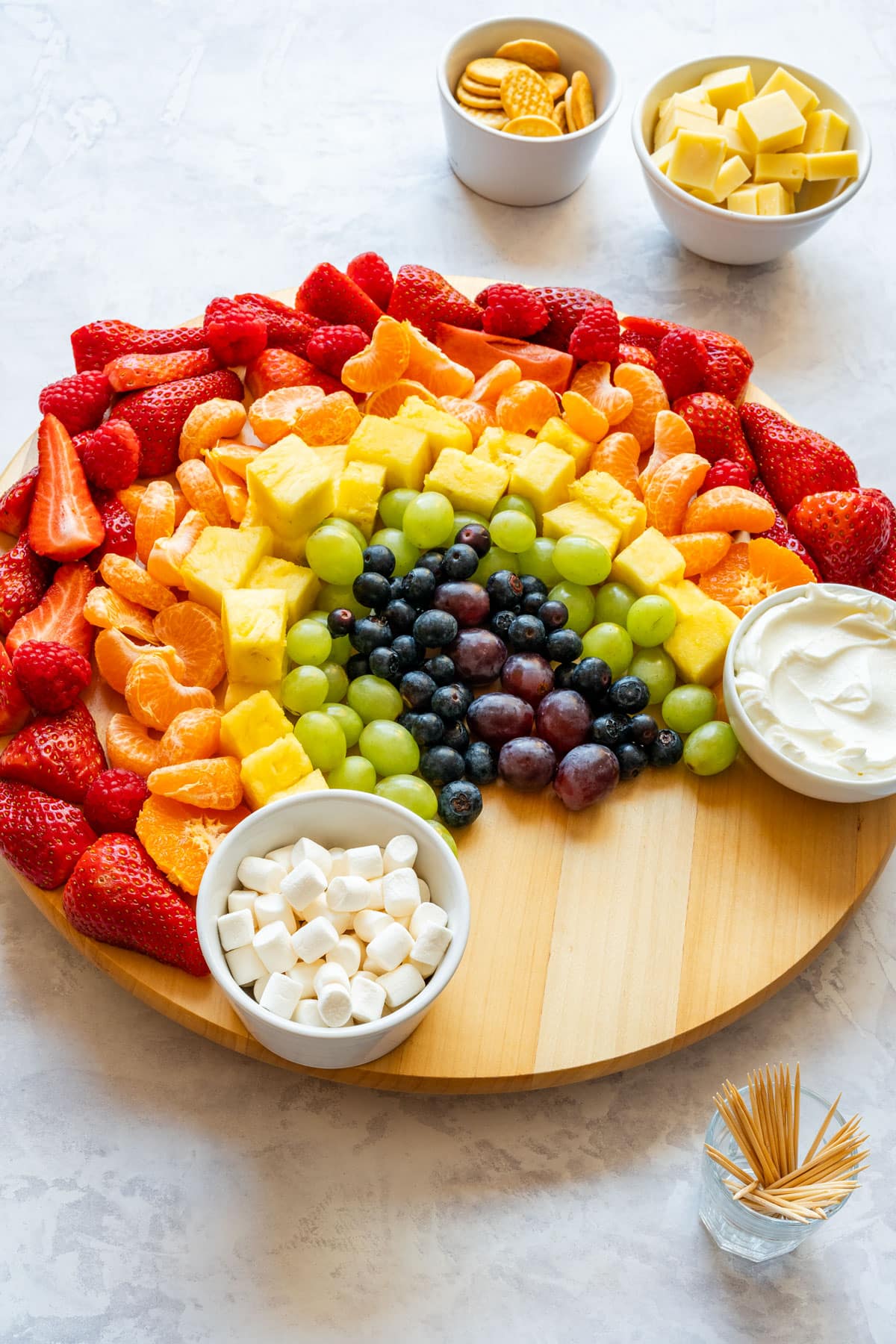 Rainbow fruit platter with fruit dip, mini marshmallows, strawberries, mandarins, pineapple, grapes and blueberries on a round board.