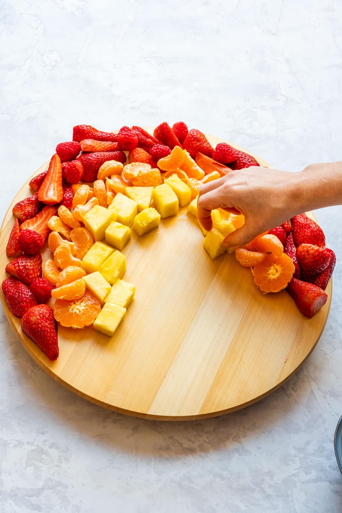 Hand adding pineapple to a fruit platter arranged in rainbow colors with strawberries, raspberries and mandarins.