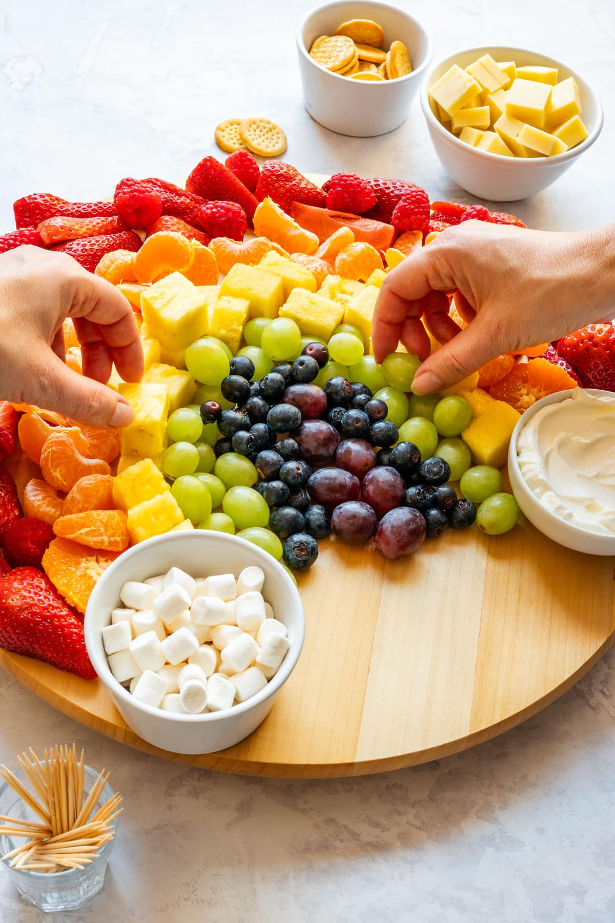 Hands reaching for a grape and a piece of pineapple from a rainbow fruit platter with fruit dip and mini marshmallows.