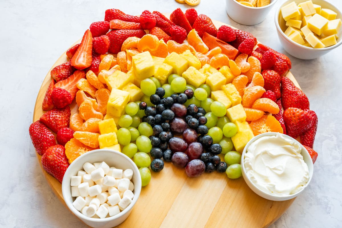 Fresh fruit arranged in rainbow colors on a round board with fruit dip and marshmallows.