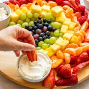 Strawberry dipped into fruit dip beside a fruit platter made with strawberries, mandarins, pineapple, grapes and blueberries.