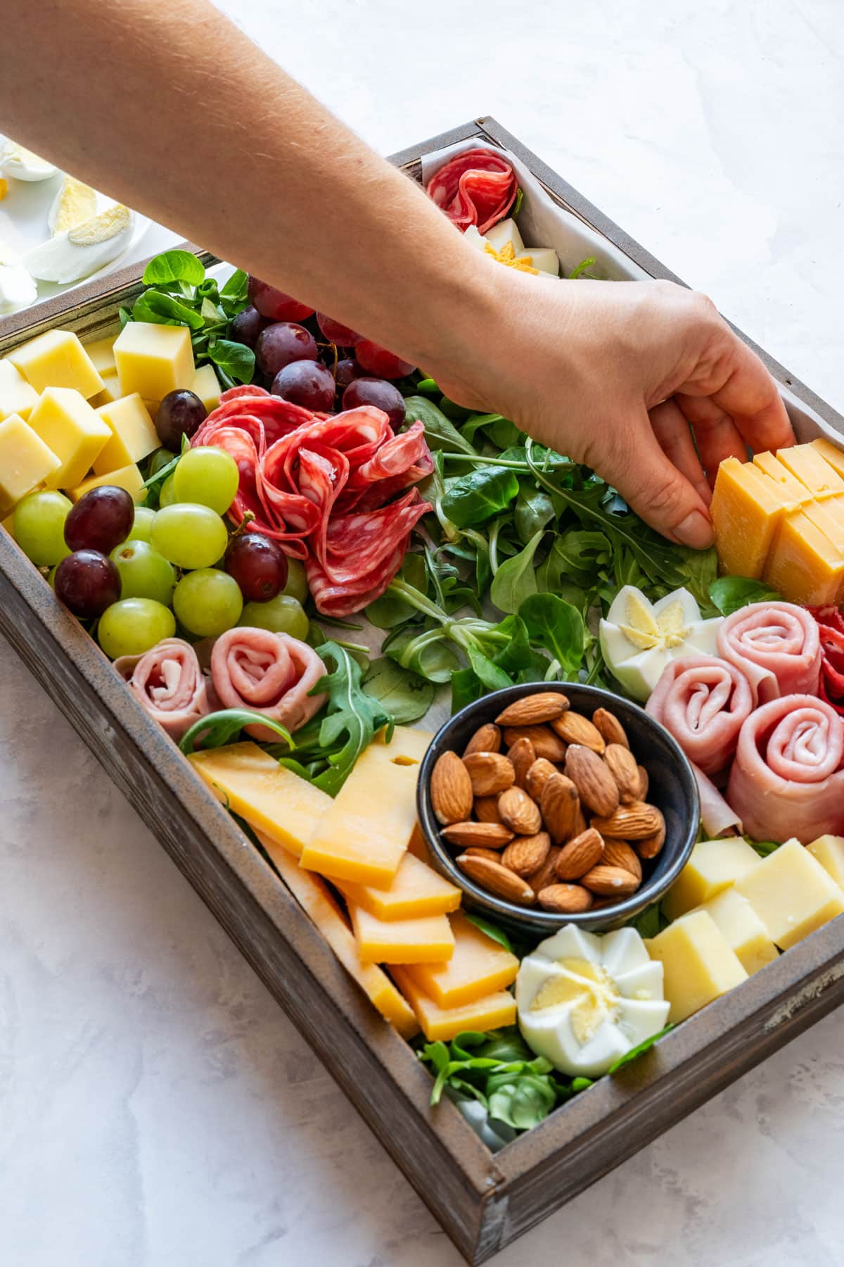 Hand placing cheese onto the board with grapes, deli meats, greens and almonds.