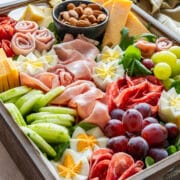 Spring charcuterie board in a wooden tray with cheese, deli meats, grapes, cucumber, almonds, crackers and sliced baguette.
