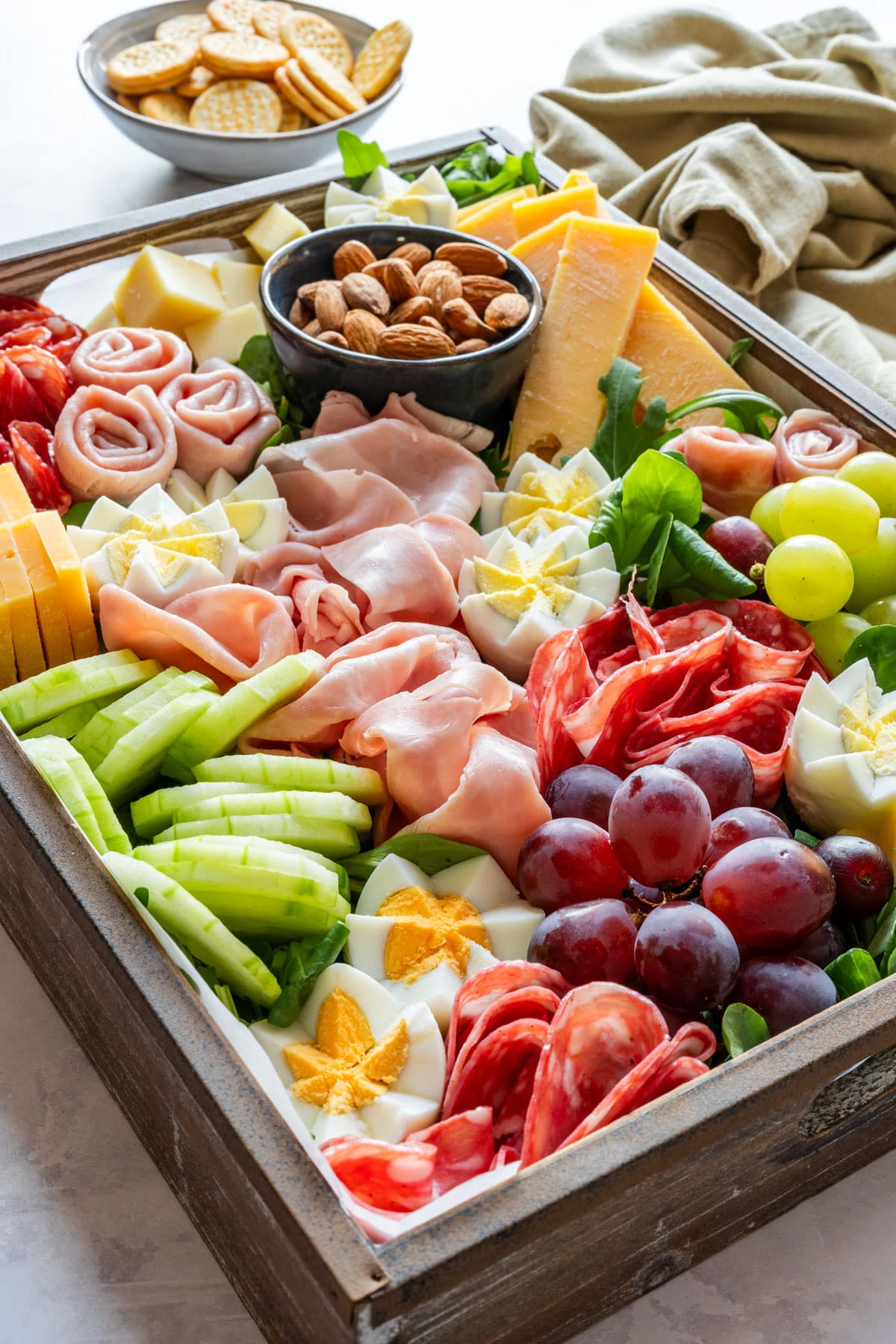 Spring charcuterie board in a wooden tray with cheese, deli meats, grapes, cucumber, almonds, crackers and sliced baguette.