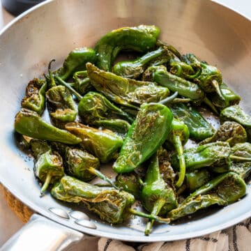 Blistered Padrón peppers with sea salt in a stainless steel pan, with lemon wedges and a bowl of coarse salt.